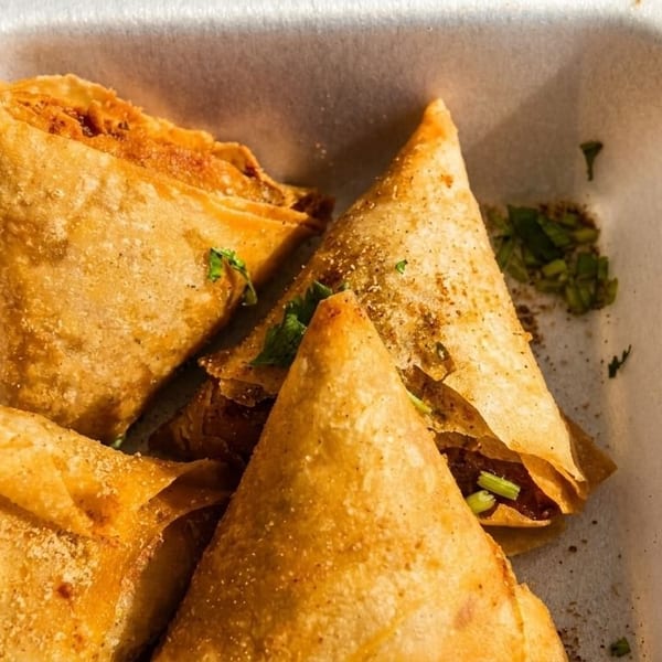 Golden-brown samosas, sprinkled with spices and herbs, in a white takeout container.