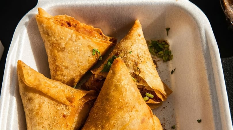 Golden-brown samosas, sprinkled with spices and herbs, in a white takeout container.