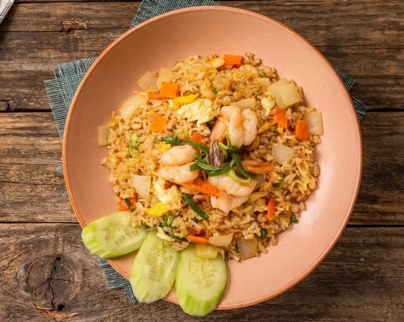 Shrimp fried rice with vegetables and egg, garnished with cucumber slices on a pink plate.