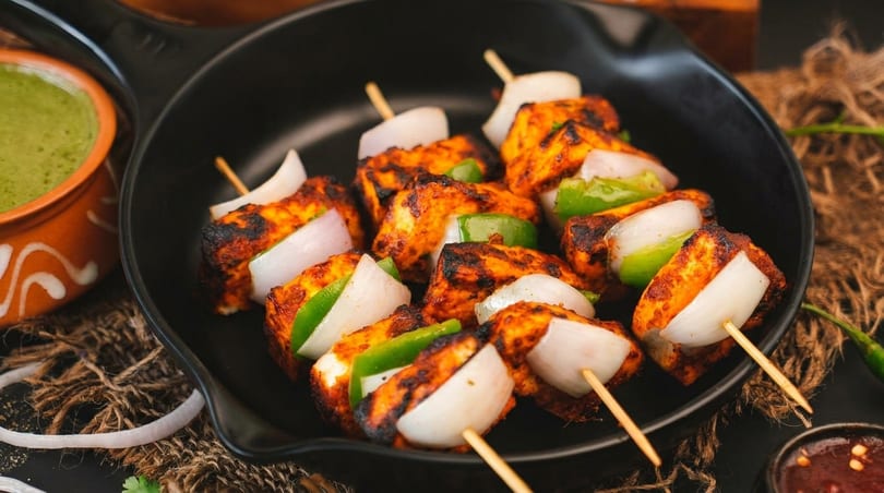 Paneer tikka skewers with charred paneer, green bell peppers, and onions in a black pan.
