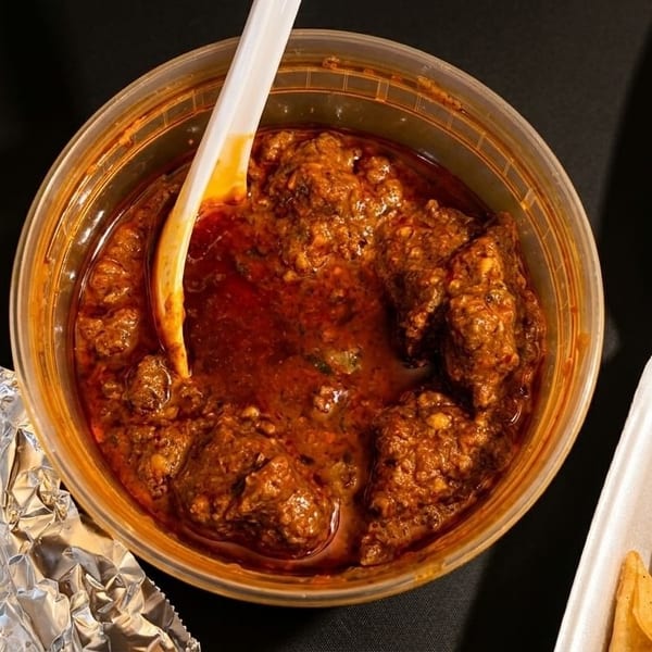 Spicy red curry with meat chunks and a white spoon in a clear plastic container.