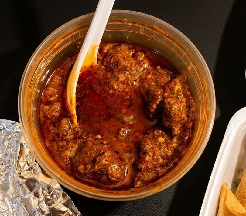 Spicy red curry with meat chunks and a white spoon in a clear plastic container.