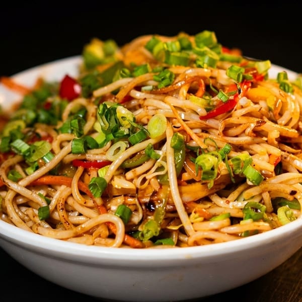 Close-up of a white bowl filled with stir-fried noodles, vegetables, and green onions.